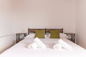 Un lit bien fait, avec du linge de lit blanc et deux coussins verts, flanqué de deux tables de chevet sur lesquelles sont posées des lampes.