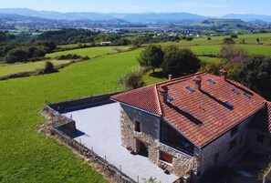 A stone building with a red-tiled roof, surrounded by green fields and distant mountains.