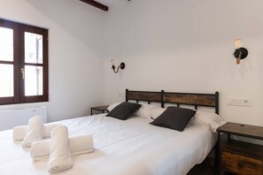 A neatly made bed with white linens and two pillows, a bedside table with a lamp, and a window with a view of the outside.