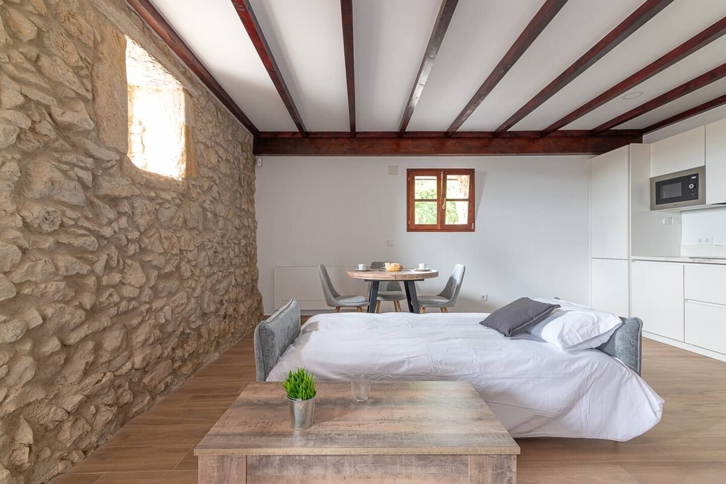 A bedroom with a stone wall, a bed with grey bedding, a wooden table with a plant, and a small kitchen area with a microwave.