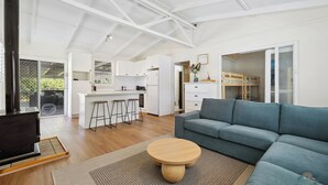 Amplia sala de estar con un sofá y una mesa redonda de madera, una kitchenette con gabinetes blancos y un ventanal con vistas a zonas verdes.