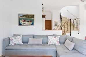 Ein Wohnzimmer mit grauem Sofa, floralen Kissen, einem Gemälde eines Bootes, einer Treppe und einer Holztür.