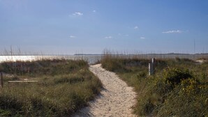 Un sendero arenoso que conduce a una playa con vegetación escasa y un cielo despejado.