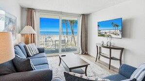 A living room with a sofa, a coffee table, and a TV on a wall. There is a balcony with a view of the beach and palm trees. - New Reno Gulf-Front 3 Bedroom Condo | Heated Pool & Hot Tub | Sand Castle I (Indian Shores)