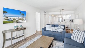 A living room with a blue sofa, a coffee table, and a TV displaying a beach scene. - New Reno Gulf-Front 3 Bedroom Condo | Heated Pool & Hot Tub | Sand Castle I (Indian Shores)