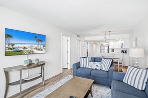 A living room with a blue sofa, a coffee table, and a TV displaying a beach scene. - New Reno Gulf-Front 3 Bedroom Condo | Heated Pool & Hot Tub | Sand Castle I (Indian Shores)