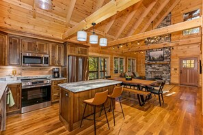 A spacious kitchen with wooden cabinets, a large island with bar stools, a dining area with a table and chairs, and a stone fireplace. - New build cabin Hocking Hills | Hot tub| Sleeps 10 (Logan)