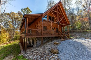 Eine Holzhütte mit Steinfundament, einem Balkon und einer Schotterzufahrt, umgeben von Bäumen.