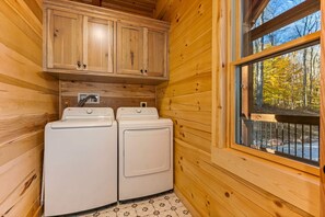 Eine gemütliche Holz-Blockhütte mit zwei weißen Waschmaschinen, Holzschränken und einem Fenster mit Blick auf einen Wald.