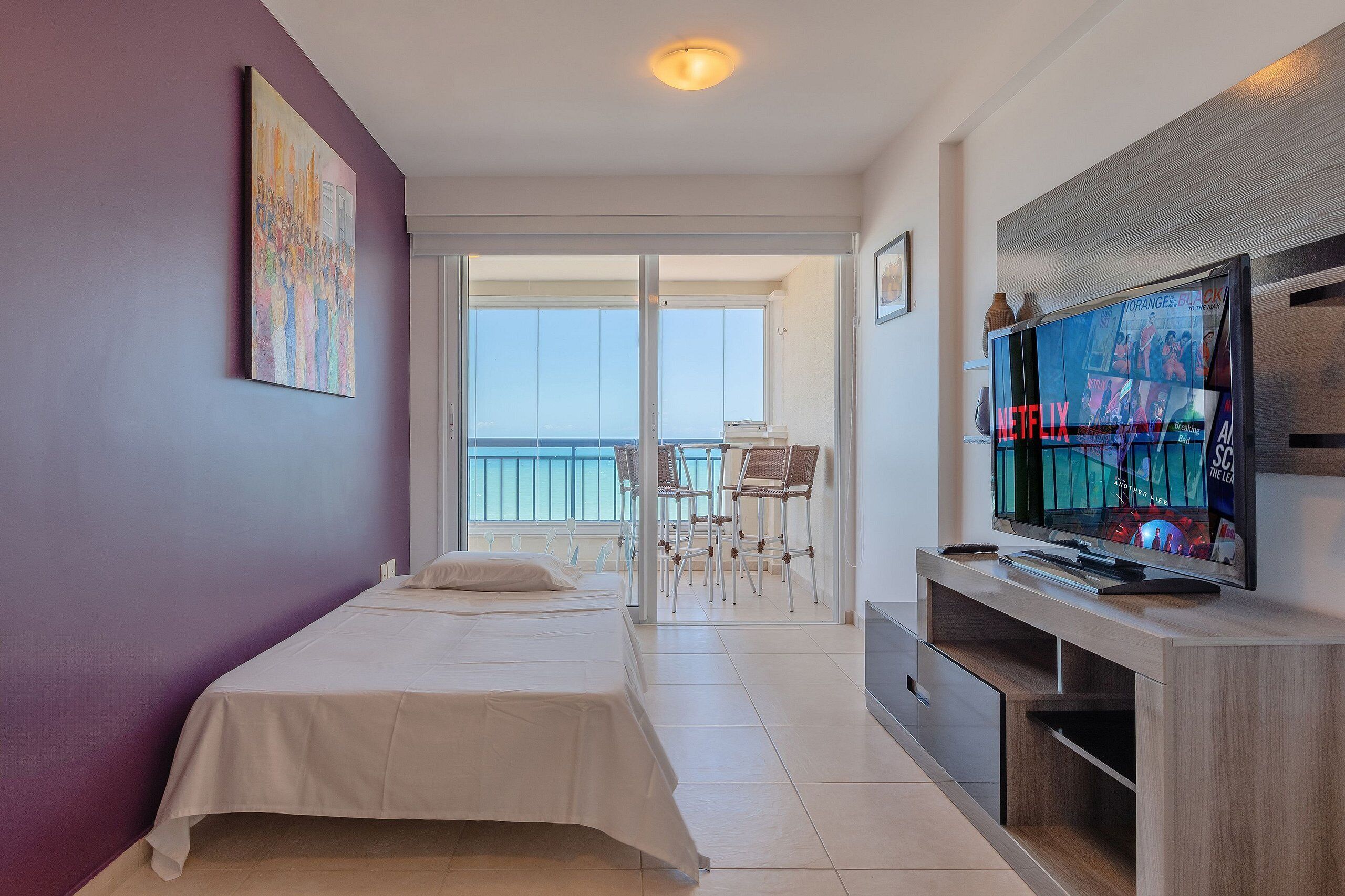 A hotel room with a bed, a TV, a balcony with chairs, and a view of the sea.