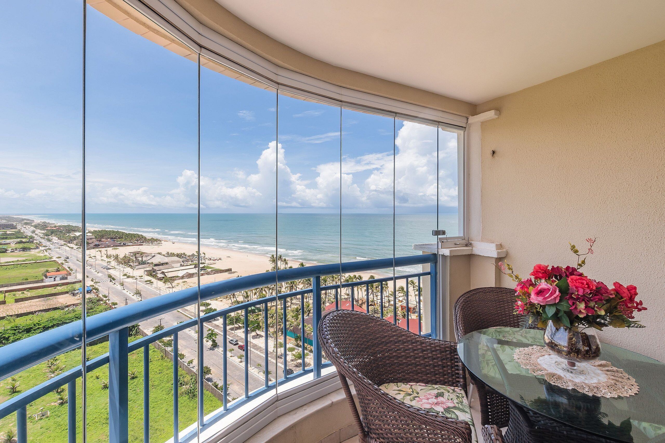 Varanda com vista para a praia e oceano, uma cadeira de vime, uma mesa de vidro com um vaso de flores e um corrimão azul.