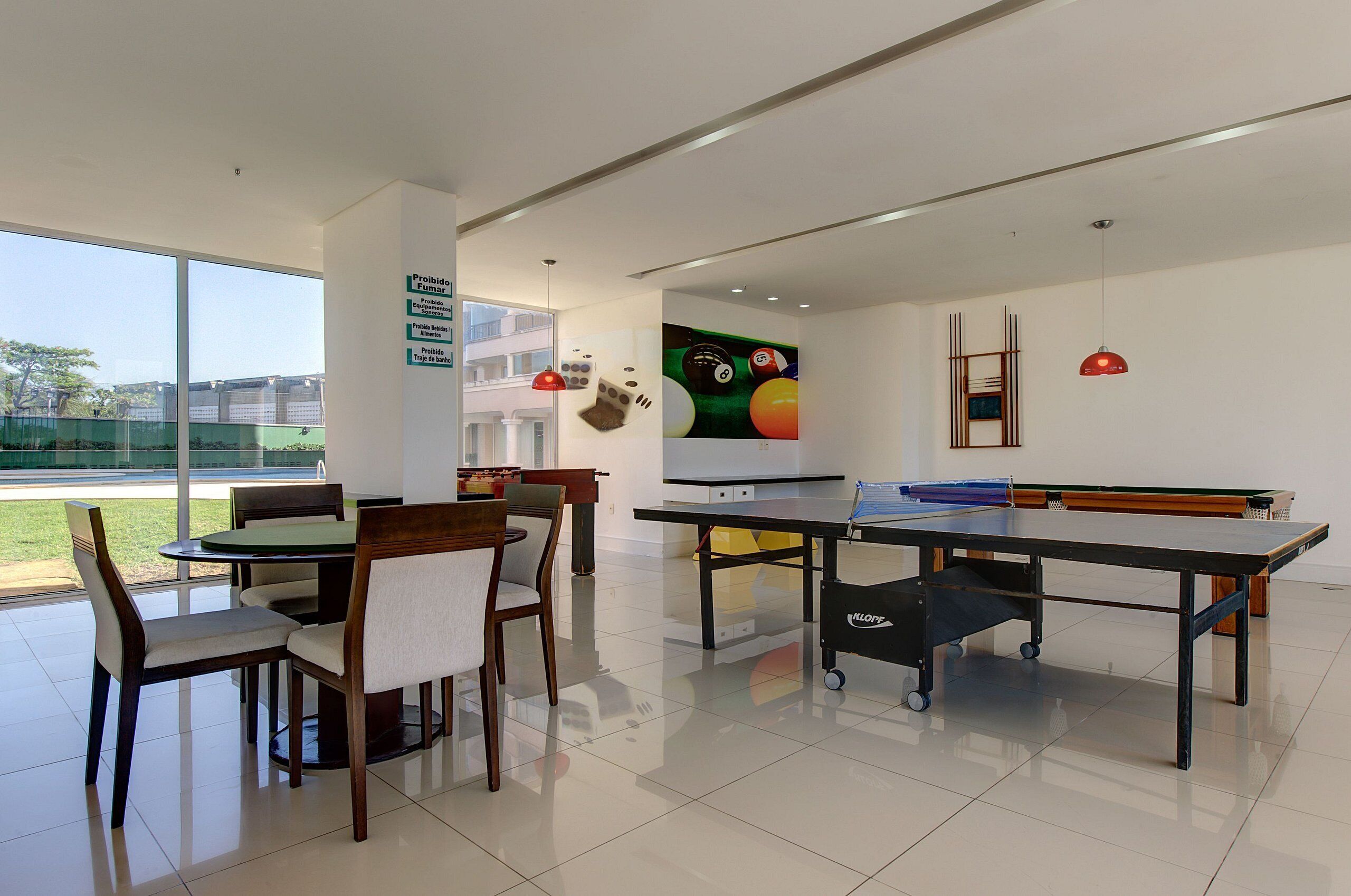 A modern indoor space with a billiards table, a dining area with chairs and a table, and a large window offering a view of a tennis court.