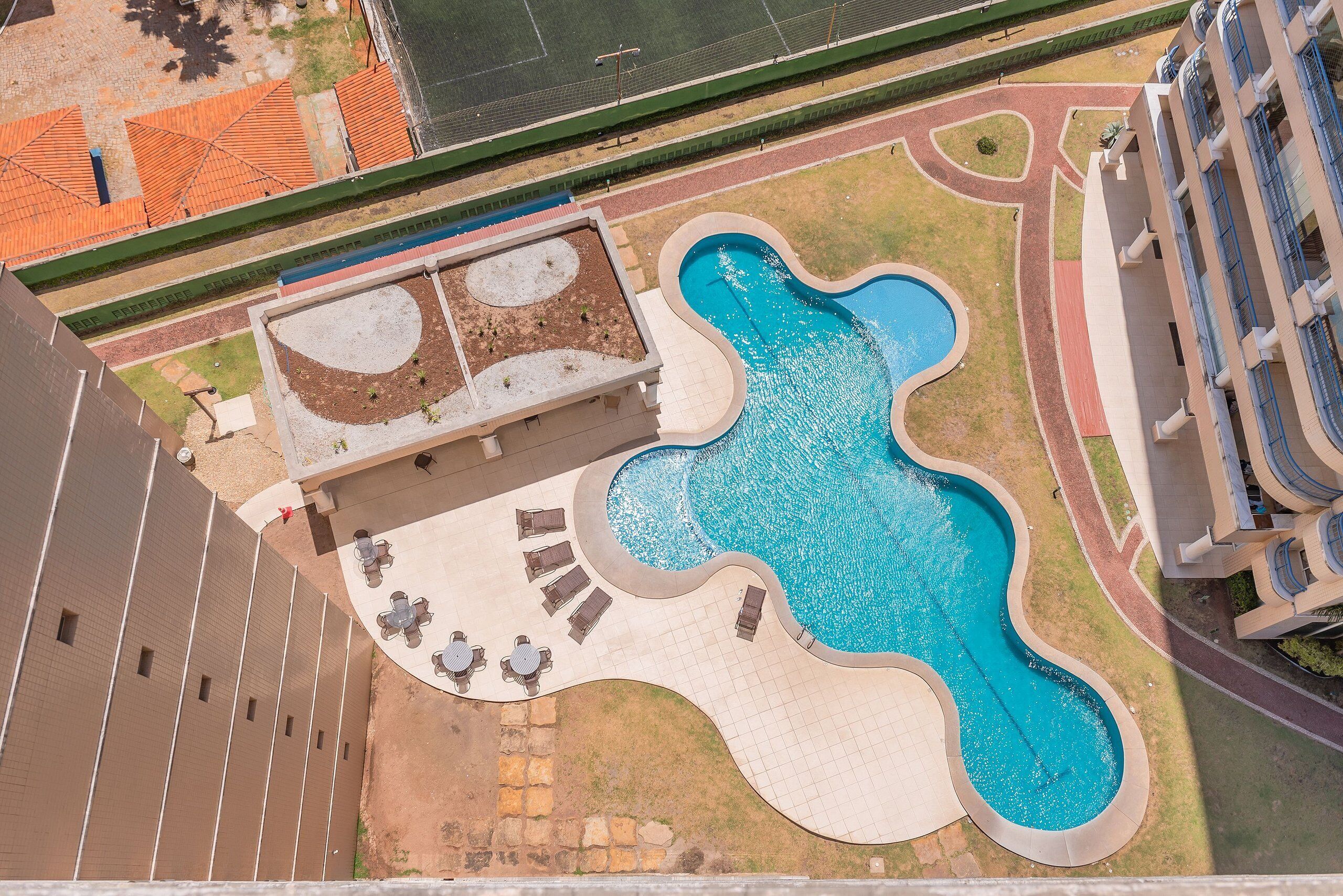 Vista aérea de uma piscina com um design ondulado singular, cercada por espreguiçadeiras e uma pequena área ajardinada.
