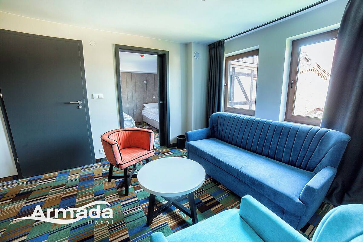 Una habitación de hotel con un sofá azul, un sillón rojo, una mesita blanca pequeña y una alfombra estampada.