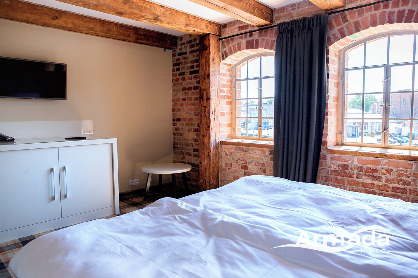 Quarto de hotel com uma cama, uma mesa redonda pequena, uma televisão fixada na parede e uma janela com cortinas.