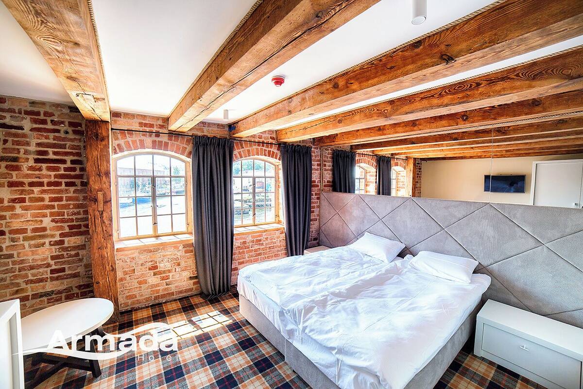 Un dormitorio con pared de ladrillo, vigas de madera, una cama grande, un escritorio y una ventana con cortinas.