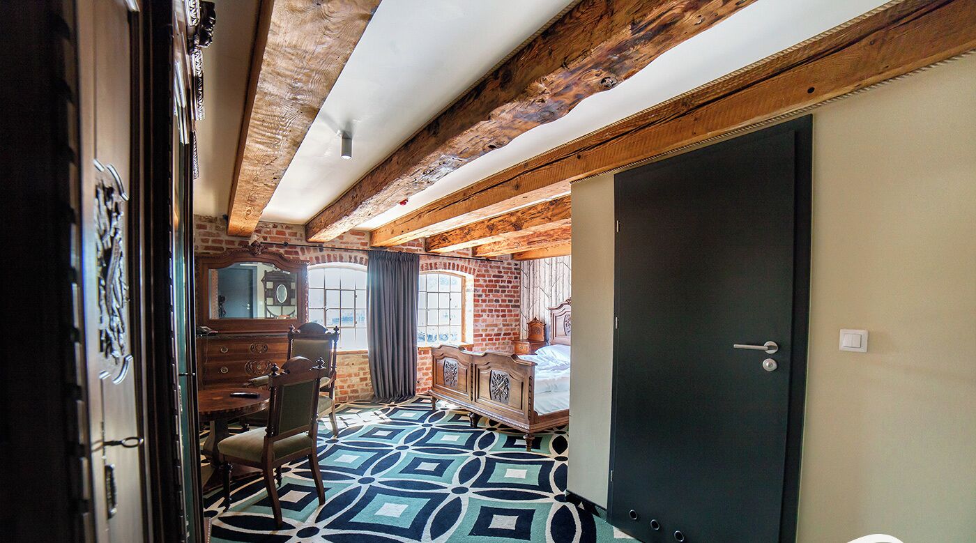 A room with exposed wooden beams, a patterned floor, a bed, a dresser, and a door.