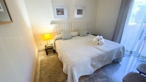 A hotel room with a large bed, a nightstand with a lamp, two framed pictures on the wall, and a view of the outdoors through the window. - Elegant Family Apartment in Aloha Golf Marbella (Marbella)