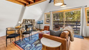 Un salon chaleureux avec une cheminée en pierre, des poutres apparentes en bois et une grande fenêtre donnant sur une terrasse.