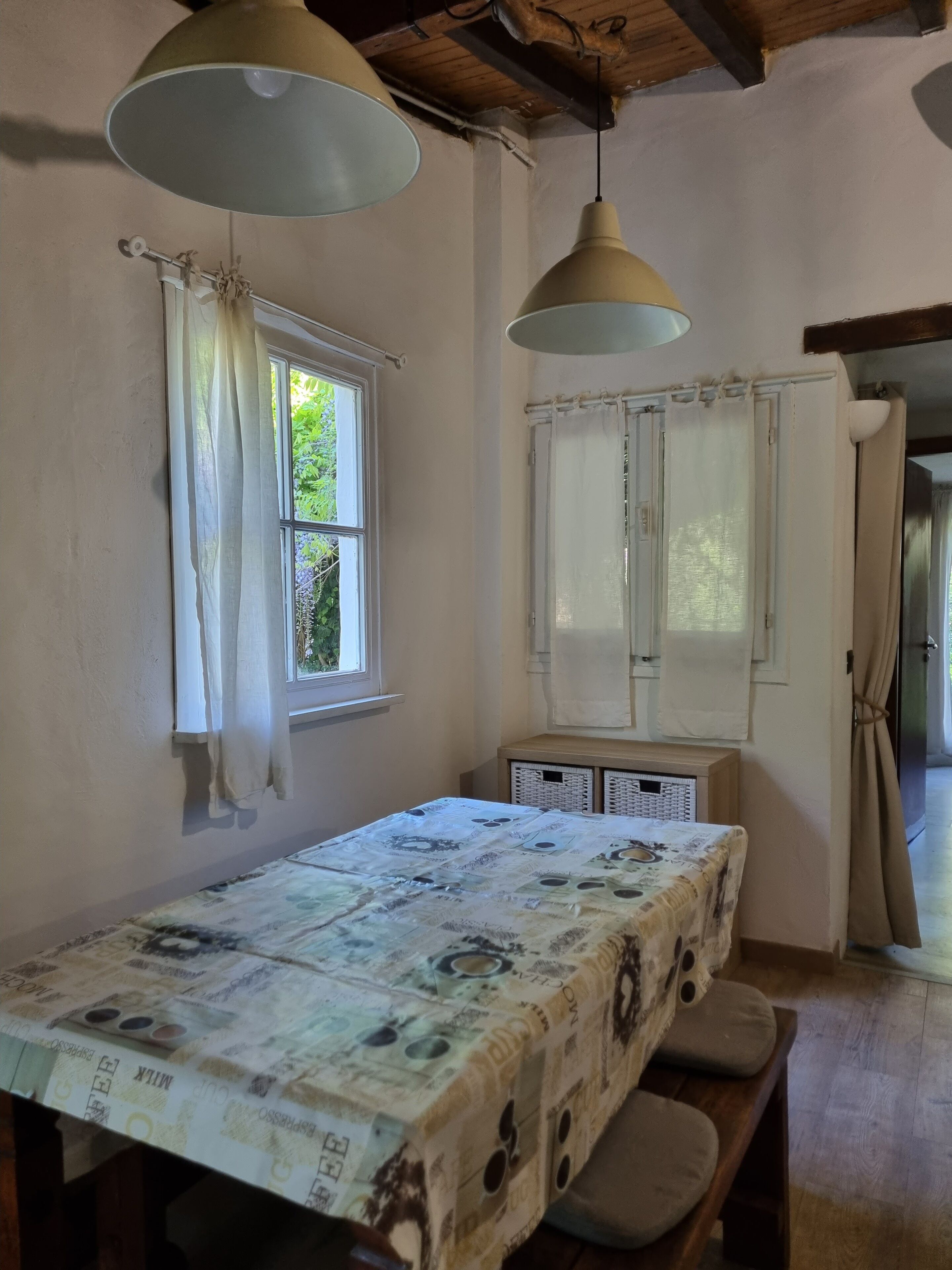 A room with a bed, a table with a patterned tablecloth, two hanging lamps, and a window with curtains.