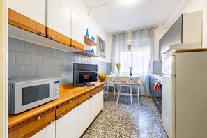 Eine kompakte Küche mit Mikrowelle, Backofen und Kühlschrank. Ein Esstisch mit Stühlen und ein Fenster mit Vorhängen sind vorhanden.