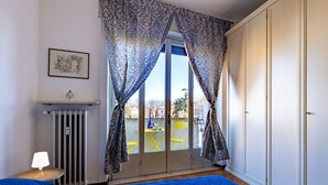 Une chambre à coucher avec un lit, un radiateur, une fenêtre avec des rideaux, un tableau encadré au mur et une vue sur l’extérieur.