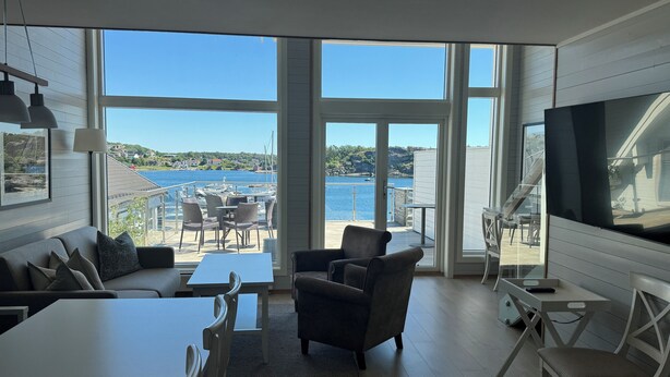 A modern living room with a large TV, a sofa, a dining table, and a view of a marina. - Havna Tjøme Apartments  (Faerder)