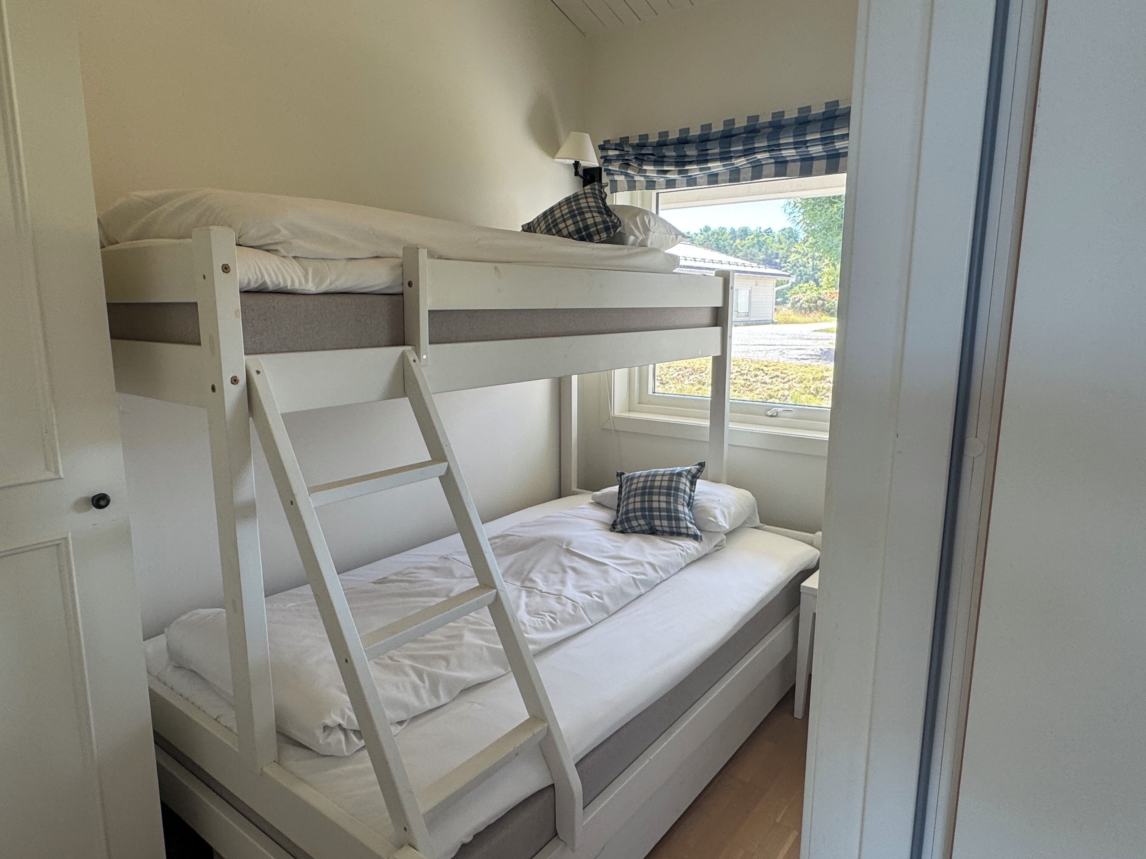 Quarto com beliche, uma janela com cortina xadrez e um abajur no parapeito.