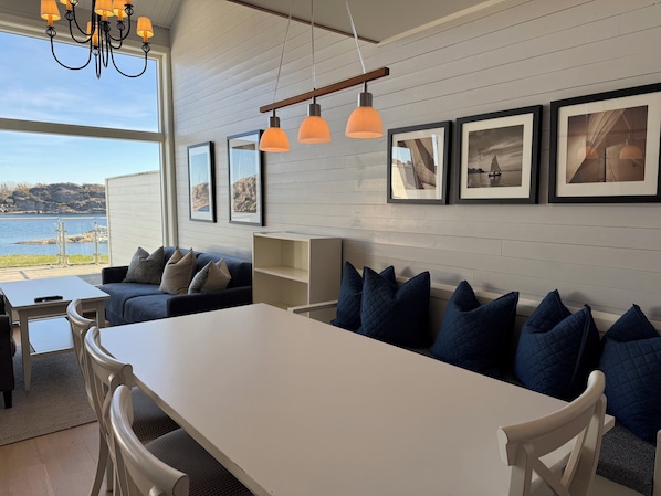 A modern living room with a large window overlooking a body of water, a dining area with a white table, and a sofa with blue cushions. - Havna Tjøme Apartments  (Faerder)