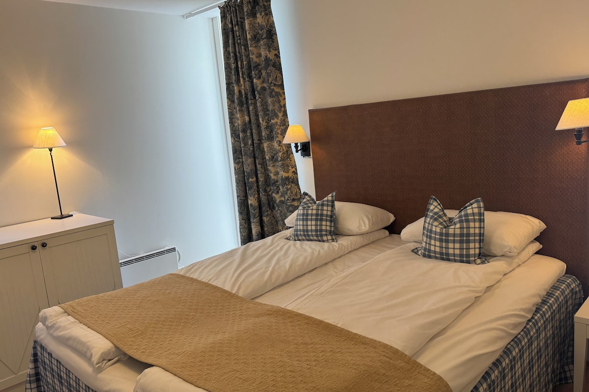 Ett hotellrum med en säng, ett nattduksbord med en lampa, ett fönster med gardiner och en sänggavel.