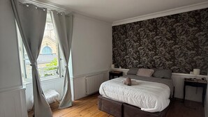 Un dormitorio con una cama grande, piso de madera, una ventana con cortinas y un papel tapiz floral en la pared.