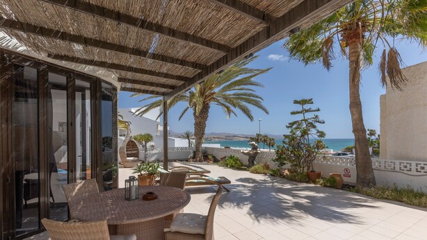 Een terras buiten met rieten meubels, een tafel en stoelen, met uitzicht op een kustgebied met palmbomen en de zee.