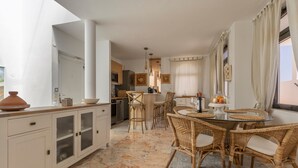 Een moderne keuken met witte kasten, een eethoek met rieten stoelen en een groot raam met gordijnen.