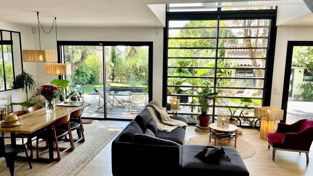 Ein modernes Wohnzimmer mit einer Couch, einem Esstisch, Stühlen, großen Fenstern mit Blick ins Grüne und einem Fahrrad im Hintergrund.