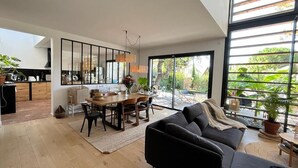 Ein modernes Wohnzimmer mit einem großen schwarzen Sofa, einem Essbereich mit Holztisch und -stühlen sowie großen Fenstern mit Blick auf Grünflächen.