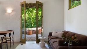 Une pièce avec un canapé marron, une petite table ronde, une chaise en bois et une porte donnant sur un espace extérieur agrémenté de plantes en pot.