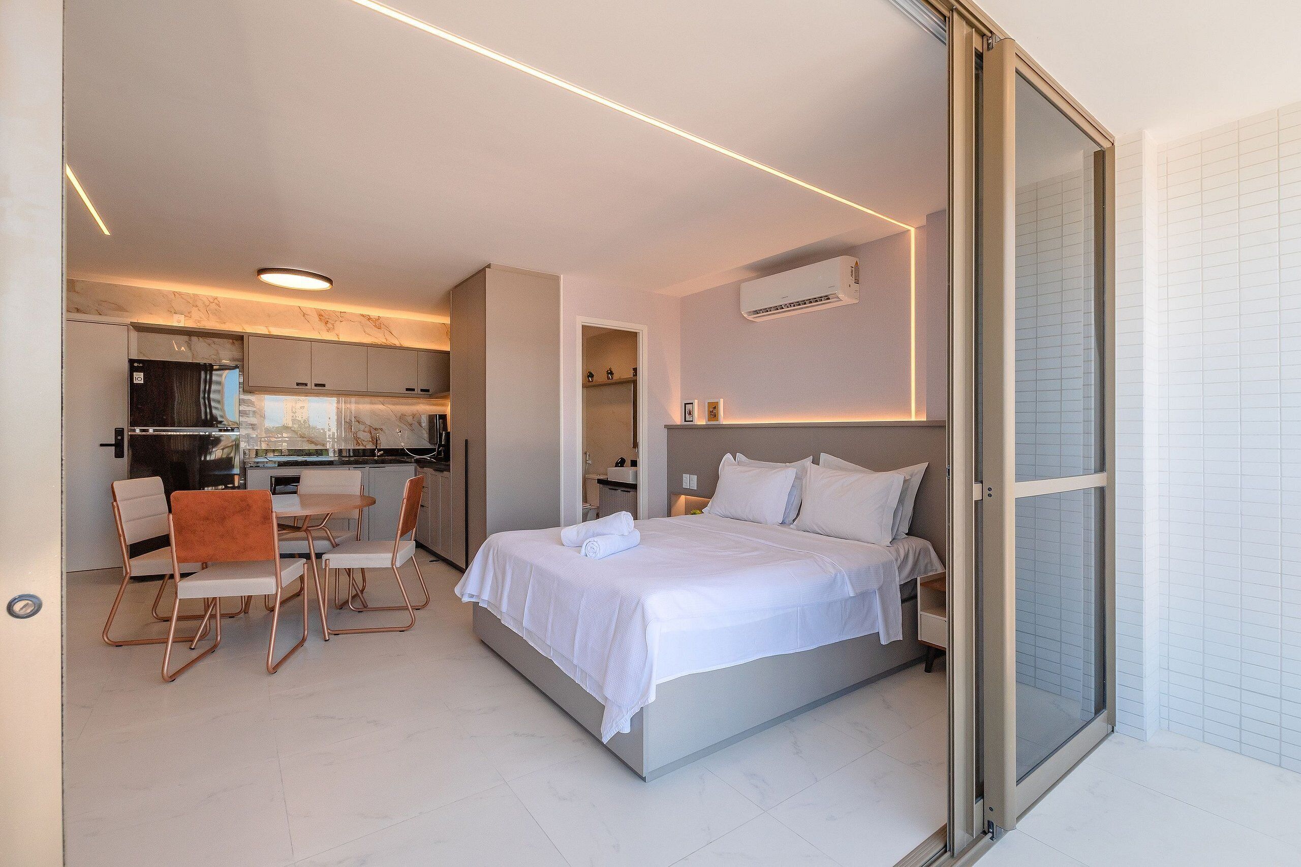 Una habitación de hotel moderna con una cama, un área de comedor con sillas, una cocina al fondo y una puerta corrediza al baño.