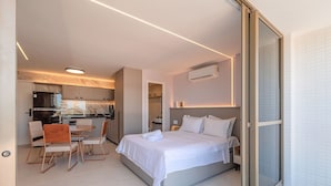 Ein modernes Hotelzimmer mit einem Bett, einer Essecke mit Stühlen, einer Küche im Hintergrund und einer Schiebetür zum Badezimmer.