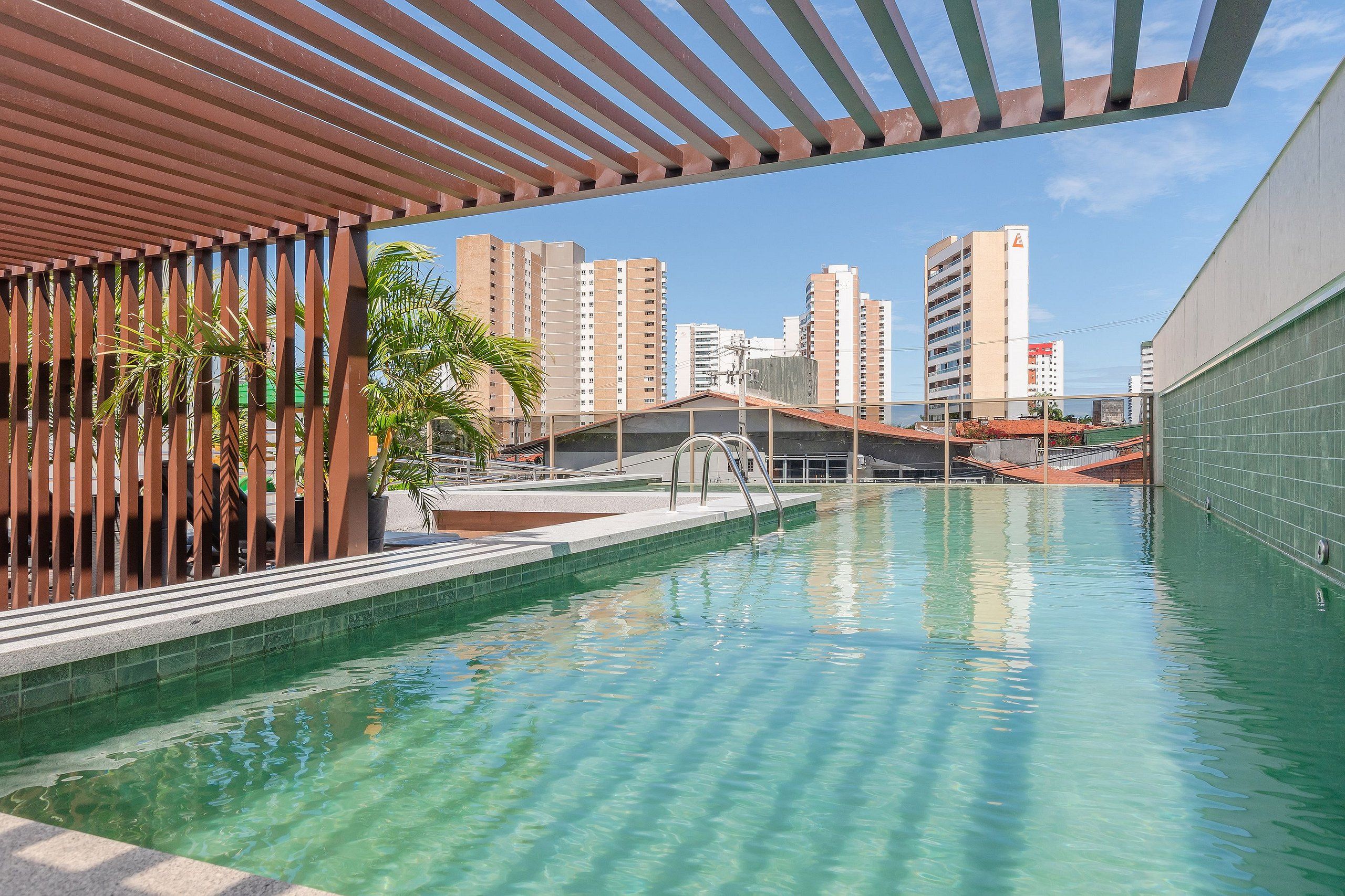 Una piscina con agua cristalina, rodeada por una estructura de pérgola y con vistas a edificios altos al fondo.