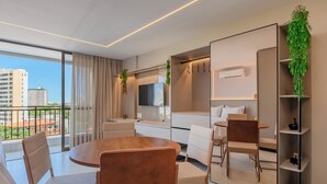 Habitación de hotel moderna con una mesa de comedor redonda de madera, sillas blancas, un espejo grande, un armario empotrado y un balcón con vistas a la ciudad.