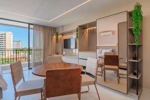 A modern hotel room with a round wooden dining table, white chairs, a large mirror, a built-in wardrobe, and a balcony with a city view.