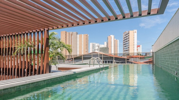 Ein Schwimmbecken mit klarem Wasser, umgeben von einer Pergola und Blick auf hohe Gebäude im Hintergrund.