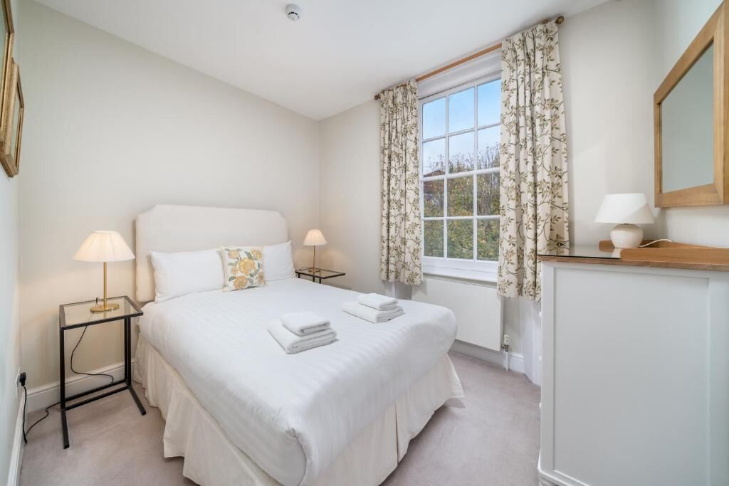 A neatly arranged bedroom with a large bed, two bedside tables with lamps, a dresser, and a window with floral curtains.