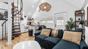 En modern lägenhet i loftstil med kök, matplats och vardagsrum. Den har en spiraltrappa, ett stort fönster och en hängande lampa.