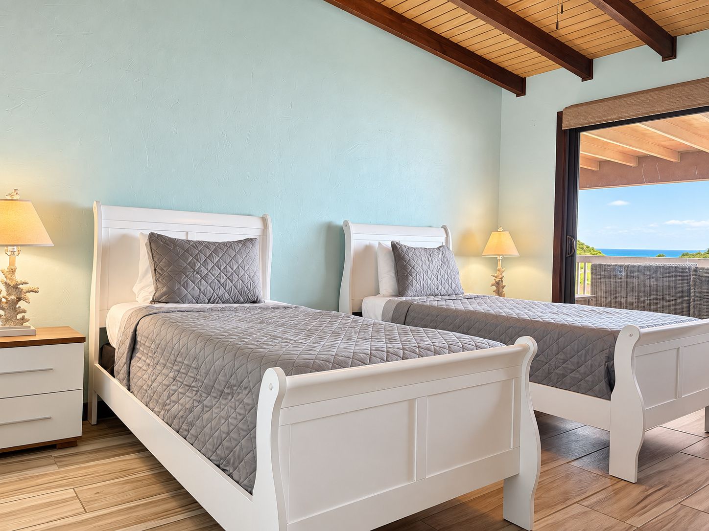 A bedroom with two beds, a nightstand, a lamp, and a view of the ocean.