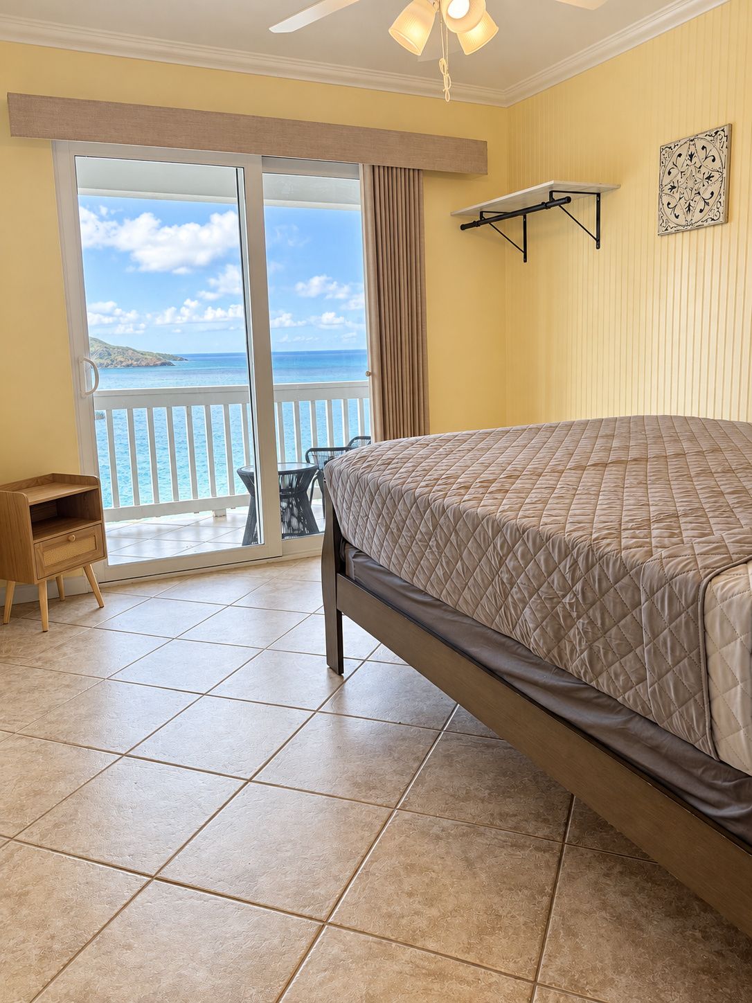 A bedroom with a bed, a small wooden cabinet, a ceiling fan, a view of the ocean, and a balcony with a chair.