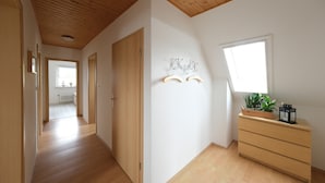 Een hal met houten vloer, houten plafond, houten deur, een witte muur met een raam, en een houten dressoir met een plant en een lantaarn.