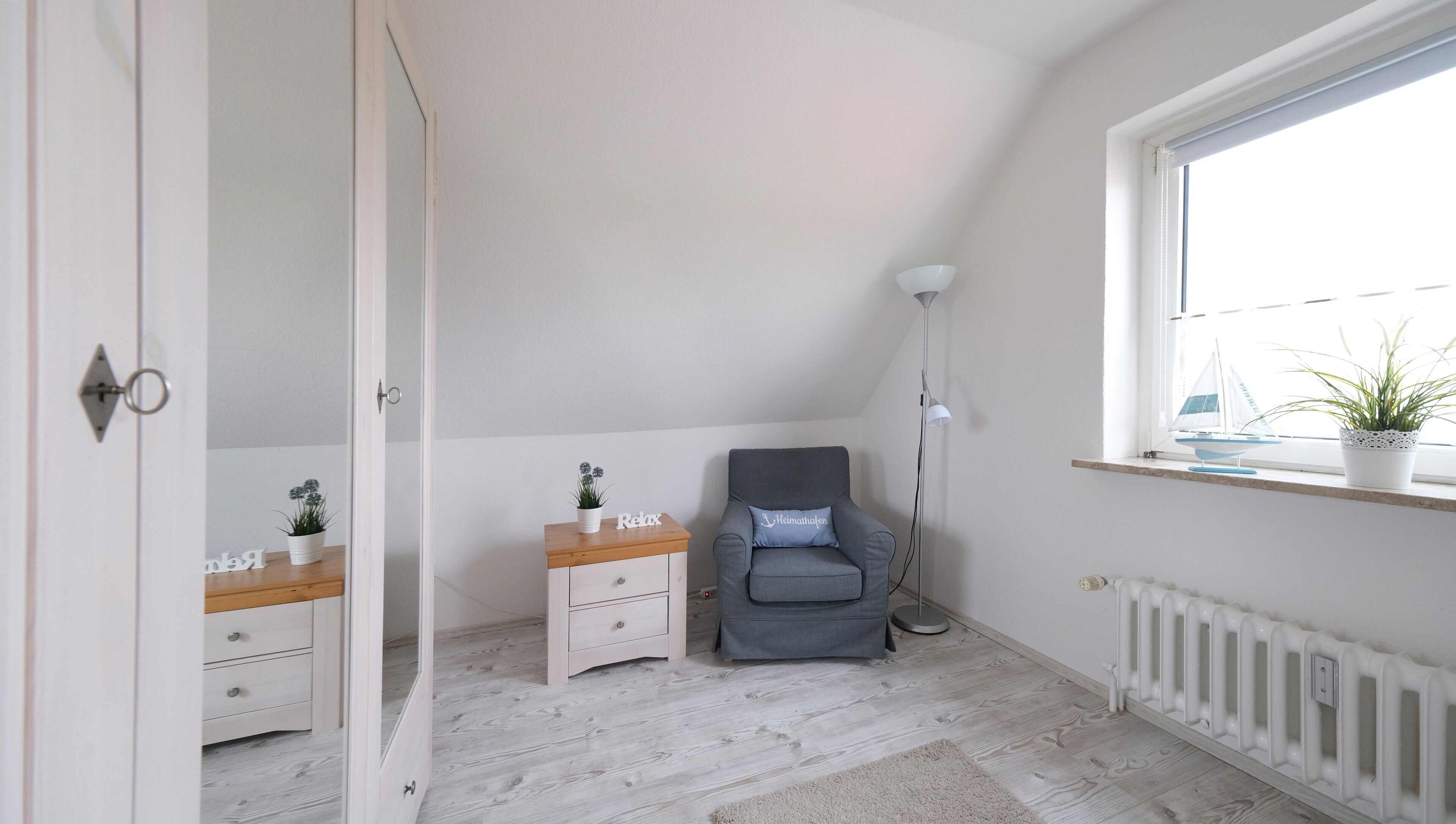 Una habitación con suelo de madera, un sillón gris, una mesita de noche con una planta, un estante pequeño con una planta y una ventana con vista a un velero.