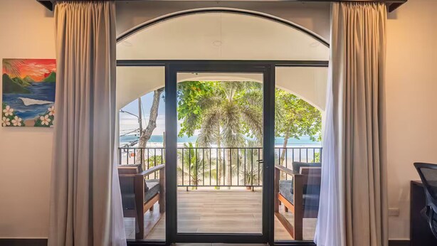 Habitación con puerta corrediza de vidrio que da a un balcón con terraza de madera, mobiliario exterior y vista a la playa y palmeras.