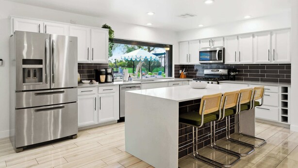 A modern kitchen with white cabinets, stainless steel appliances, a large island with bar stools, and a view of greenery outside. - Fun-Filled Oasis • Pool, Pickleball, Games Galore (Scottsdale)